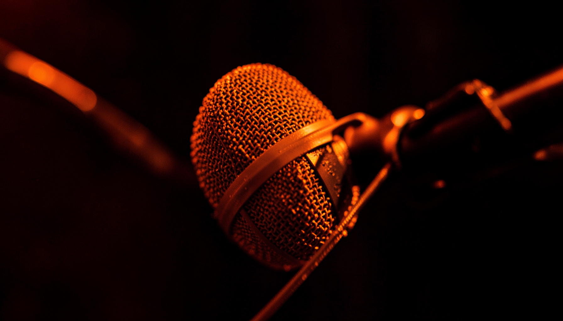 Close-up of a studio microphone on a boom arm against a dark blurred background, warm orange accent light