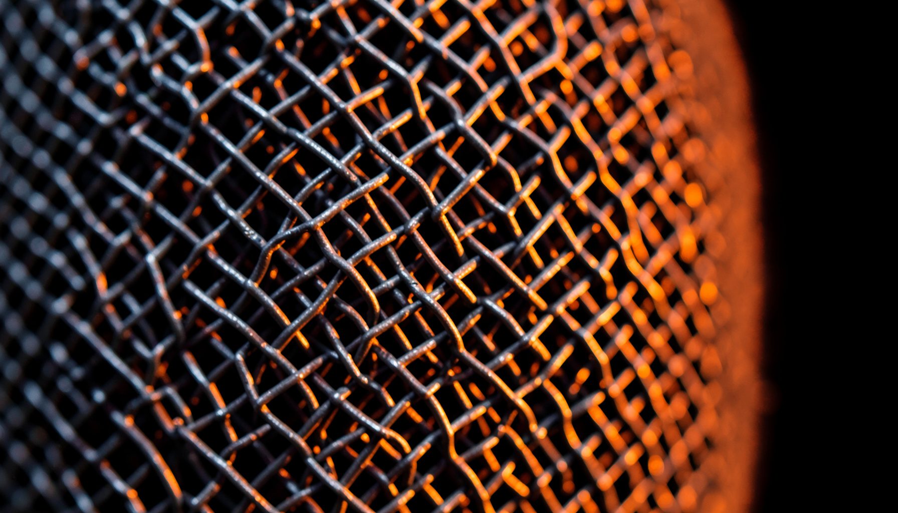 Extreme close-up of a dark gray microphone grille mesh, metallic weave catching warm amber light against a black background