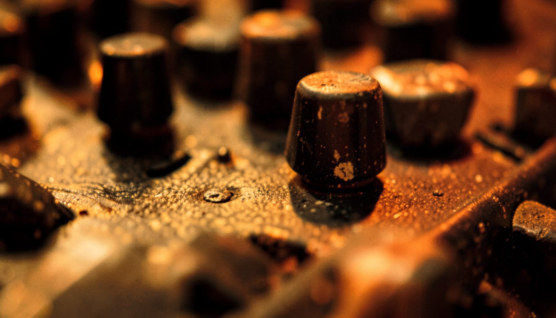 Extreme close-up of worn soundboard knobs and faders in warm amber and charcoal tones, textured and moody
